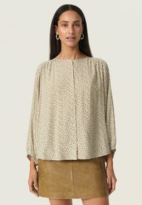Blusa beige con pois, collo tondo, chiusura con bottoni e maniche a palloncino. Abbinata a una minigonna in pelle marrone.