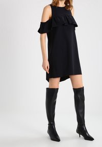 Woman wearing a black cold-shoulder dress with ruffles paired with black knee-high heeled boots on a white background.