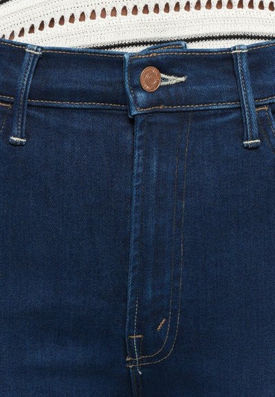 Close-up van de tailleband van donkerblauwe denim jeans met een koperen knoop, riemlussen en een deel van een witte gebreide top erboven.