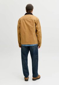 Mand iført en brun jakke, blå jeans, lysebrune støvler og en mørk beanie, stående med ryggen til mod en ensartet lys baggrund.