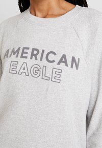 Felpa grigia con scollo a girocollo e maniche raglan. Presenta la scritta "AMERICAN EAGLE" stampata in grassetto nero e grigio sul petto.