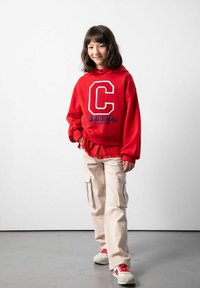 Czerwona bluza z kapturem z dużą białą literą "C" i napisem "ORIGINAL", połączona z beżowymi spodniami cargo z bocznymi kieszeniami i czerwono-białymi trampkami.