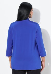 Top lungo blu royal con maniche lunghe e collo alto, caratterizzato da una texture liscia e una vestibilità comoda. Mostrato di spalle, con dettagli di cucitura semplici.