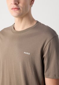 Hnedé bavlnené tričko s okrúhlym výstrihom a krátkymi rukávmi, ktoré má malé biele logo značky na ľavej strane hrude.