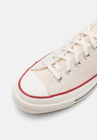Gros plan d'une basket en toile beige avec des lacets blancs, un embout en caoutchouc blanc et une bande rouge le long du bord de la semelle sur fond blanc.