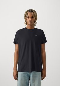 Schwarzes Kurzarm-T-Shirt mit Rundhalsausschnitt und kleinem roten Logo. Hergestellt aus glattem, leichtem Stoff, kombiniert mit hellblauen Jeans.