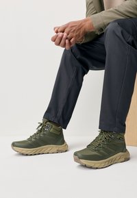 Žalios spalvos žygių batai iš patvarios medžiagos, turintys tekstūruotą paviršių, raištelių užsegimą ir tvirtą smėlio spalvos pado su sukibimo raštais dizainą.