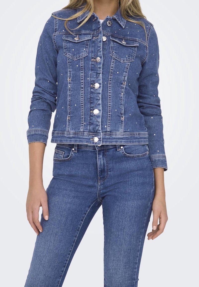 En ung kvinna modellerar en blå denimjacka med dekorativa nitar, som är parat med blå jeans, mot en ljus bakgrund.