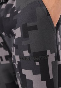 Zbliżenie ręki w kieszeni spodni w szaro-czarny cyfrowy kamuflaż z napisem "DROPSIZE" wydrukowanym na materiale.