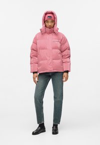 Pink puffer jacket with a shiny finish, hood, and snap closure. Paired with denim jeans and black loafers. Solid color beanie with logo.