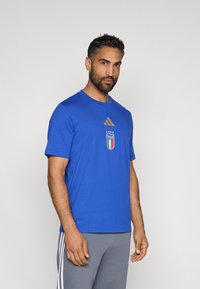 Μπλέ βαμβακερό μπλουζάκι με χρυσό λογότυπο Adidas και ιταλική εθνική σφραγίδα με τέσσερα αστέρια από πάνω. Σχεδιασμός με κοντά μανίκια και λαιμόκοψη.