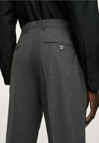 Graue taillierte Hose mit glatter Textur, schmaler Passform und zwei hinteren Taschen mit Knopfverschlüssen. Eine Kombination mit einem schwarzen Hemd ist sichtbar.