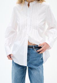 Weiße Baumwollbluse mit gefaltetem Vorderteil, Stehkragen und langen Ärmeln. Getragen mit hochgeschnittenen blauen Jeans, die eine lockere Passform zeigen.