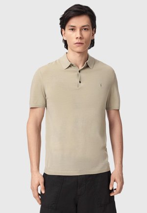 Ung mand med sort hår iført en beige kortærmet polo og sorte bukser, stående foran en ensfarvet lys baggrund.