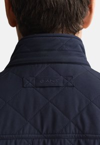 Marinblå vadderad jacka med hög krage, med ett präglat "GANT"-logo och subtilt diamantmönster, som visar en slät textur.