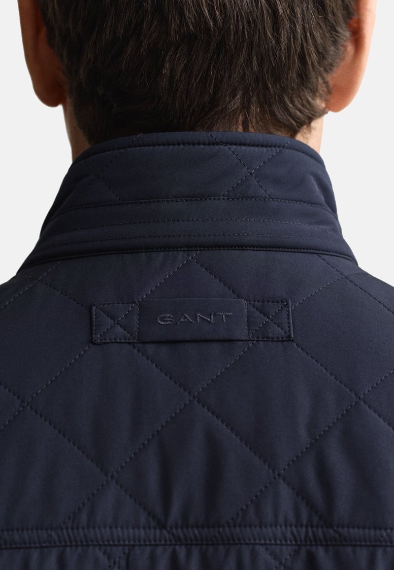 Chaqueta acolchada azul marino con cuello alto, con un logo "GANT" en relieve y un sutil patrón de diamantes, que muestra una textura suave.