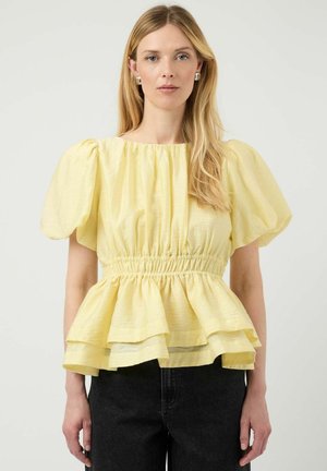 Kobieta o długich blond włosach, nosząca żółtą bluzkę z bufkami na rękawach, marszczonym stanem i warstwowym falbanowym dołem, w połączeniu z czarnymi spodniami.