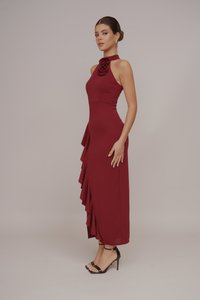 Burgundy long dress with a halter neck, featuring fabric roses at the neck, and asymmetrical ruffled hem, made from smooth material.