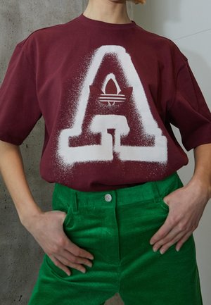 Personne portant un t-shirt bordeaux avec une grande lettre blanche taguée "A" et un pantalon en velours côtelé vert, les mains dans les poches.