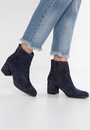 Paire de bottines en suède bleu marine à talons blocs et fermetures éclair latérales, portées avec un jean bleu clair à ourlet effiloché sur fond blanc.