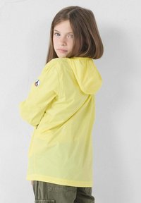 Meisje met schouderlang bruin haar, gekleed in een gele hoodie-jack en groene cargobroek, kijkt over haar schouder.