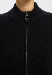 Crni, teksturirani pleteni kardigan s visokim ovratnikom i srebrnim zatvaračem. Mekan materijal, minimalistički dizajn i opuštena silueta.