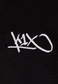 Tela negra con texto bordado en blanco que presenta un diseño estilizado "k1x", caracterizado por ángulos agudos y un final ligeramente cursivo.