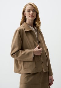 Giacca in suede sintetico marrone con una silhouette ampia, colletto grande, chiusura con bottoni e tasche frontali. Texture liscia e lunghezza leggermente accorciata.