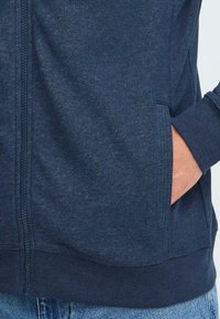 Detailný pohľad na osobu v tmavo modrom zipsovom mikine, s rukou v bočnej vrecku, v kombinácii s modrými džínami.