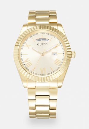 Gold-tone metal wristwatch with fluted bezel, Roman numeral markers, day and date display, and linked bracelet band by Guess.
