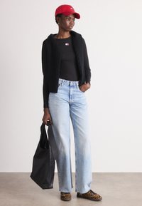 Gilet nero in maglia, top lungo a maniche nere, jeans alta vita blu chiaro, borsa nera e cappellino rosso. Scarpe con un motivo mettono in evidenza l'outfit.