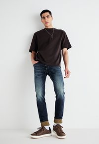 Barna oversized póló, kék slim-fit farmernadrág, barna sportcipő fehér talppal, felgöngyölt nadrágszárral; minimalista dizájn, finom fényességgel.