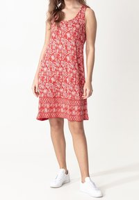 Femme portant une robe rouge sans manches avec des motifs floraux blancs et des baskets blanches, debout devant un fond blanc uni.