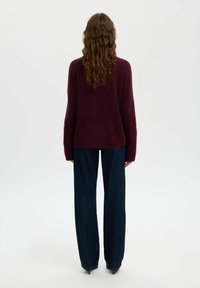 Persona con lunghi capelli ondulati, che indossa un maglione fuzzy bordeaux, jeans a gamba larga blu scuro e décolleté neri a punta, in piedi con la schiena rivolta.