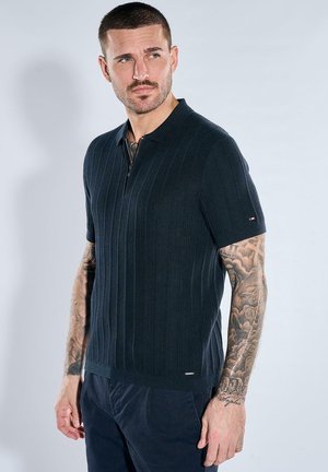 Mann mit kurzen Haaren und Tätowierungen auf beiden Armen trägt ein marineblaues, geripptes Kurzarm-Poloshirt mit Reißverschluss am Kragen.