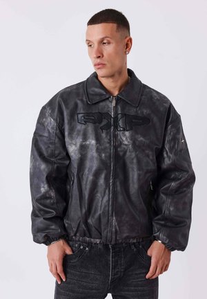 Mann trägt eine schwarze und graue abgenutzte Lederjacke mit Reißverschluss vorne und Patch-Logo, posiert mit Händen in den Taschen vor einem einfarbigen Hintergrund.