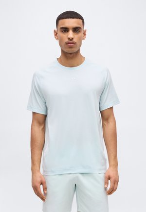 Νεαρός άνδρας με κοντά μαλλιά, που φοράει ανοιχτό μπλε κοντομάνικο μπλουζάκι και ταιριαστά σορτς, στέκεται μπροστά σε απλό φόντο.