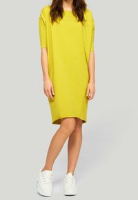 Żółta sukienka z krótkim rękawem, luźny krój, do kolan, o gładkiej fakturze. W zestawie z białymi sneakersami.