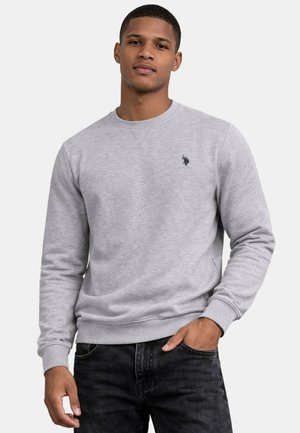 Young man wearing light gray long-sleeve sweatshirt with small embroidered logo, black jeans, standing with one hand in pocket.