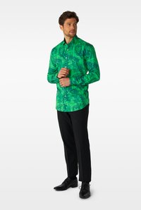 Grünes Langarmshirt mit aufwändigem Dschungelmuster, Hemdkragen mit Knöpfen und geradem Saum, kombiniert mit schwarzen Hosen und Schuhen.