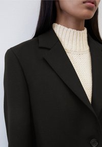 Blazer noir avec des épaules structurées, des revers en notch et un seul bouton. Porté par-dessus un pull à col roulé en maille côtelée crème. Texture lisse.