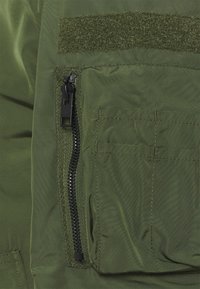 Oliwkowa kurtka z zamkiem błyskawicznym, teksturowana tkanina, miejsce na rzep, oraz płaskie detale kieszeni, które podkreślają cechy funkcjonalnego designu.