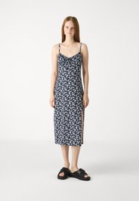 Marineblå blomster-midi kjole med tynne stropper, sideslitt og knytedetalj ved halsen. Myk stofftekstur, tettsittende silhuett. Svarte sandaler.