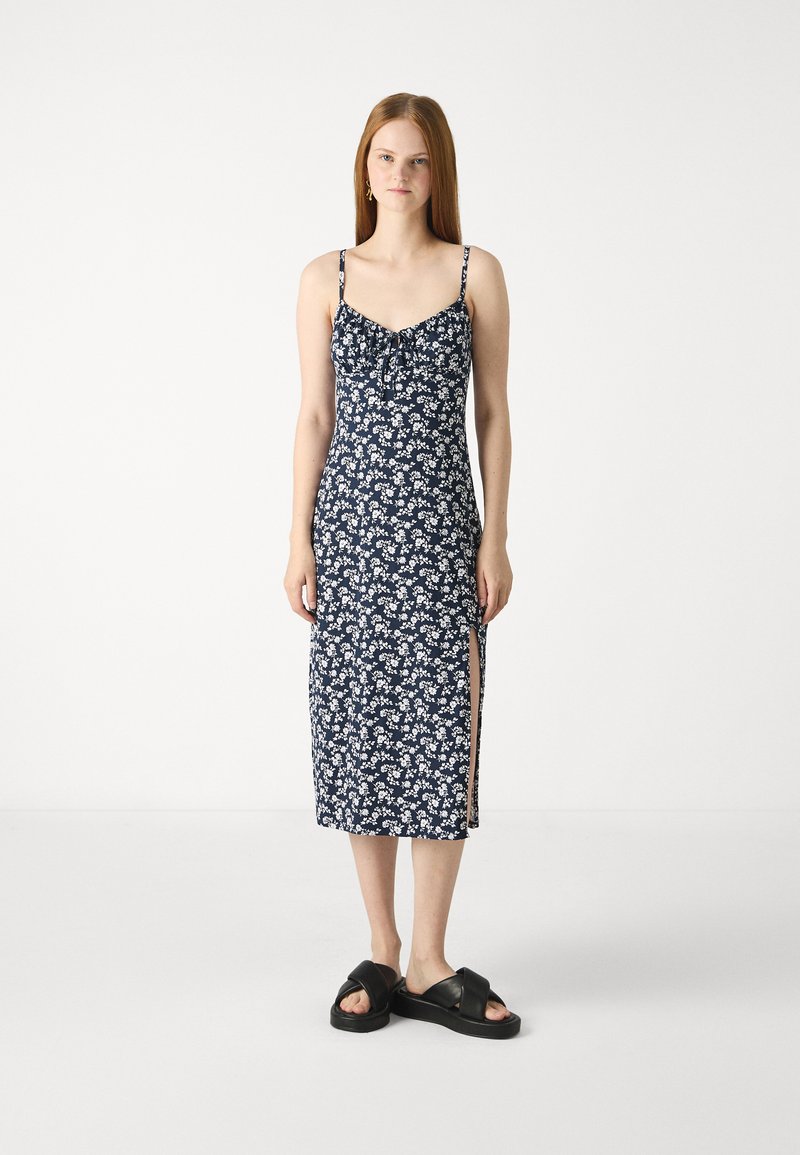 Marineblå blomster-midi kjole med tynne stropper, sideslitt og knytedetalj ved halsen. Myk stofftekstur, tettsittende silhuett. Svarte sandaler.