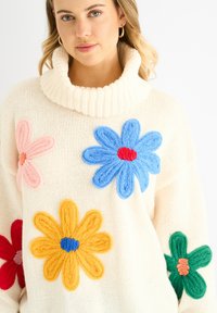 Pullover in crema con colletto alto a coste. Presenta grandi applicazioni floreali colorate in rosa, blu, giallo e verde.