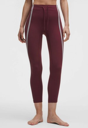 Vrouw draagt bordeauxrode leggings met hoge taille, witte zijstrepen en een trekkoord in de taille, staat op blote voeten tegen een witte achtergrond.