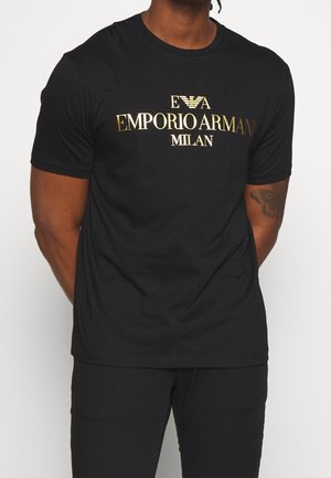 Mężczyzna ubrany w czarną koszulkę Emporio Armani Milan z złotym logo, stoi z rękami za plecami, z widocznym tatuażem na prawej ręce.