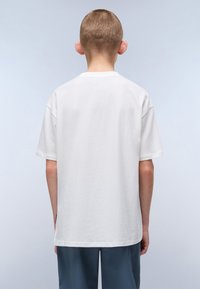 Bílá bavlněná tričko s krátkými rukávy a rovný střih. Má kulatý výstřih a minimalistický design, nošeno s světle modrými kalhotami.