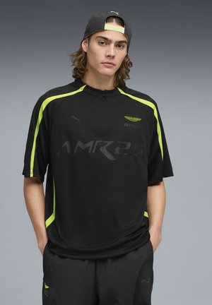 Junger Mann mit langen Haaren, der ein schwarzes und neongelbes Sportshirt sowie passende Hosen trägt, die Hände in den Taschen hat und eine rückwärts aufgesetzte Kappe trägt.