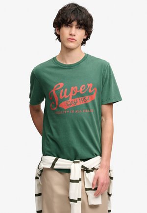 Ung mand med mørkt krøllet hår iført en grøn T-shirt med rød tekst "Super Dry 1954" og en hvid sweater bundet om beige bukser.